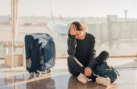 verzweifelte Frau, die ihren Anschlussflug verpasst hat, sitzt mit einem blauen Koffer auf dem Boden am Gate und hat ihr Gesicht in ihrer Hand vergraben